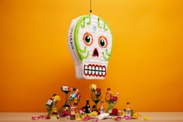A colorful skull-shaped piñata hangs against an orange backdrop. Below it is a wooden surface with various small liquor bottles, roses, and candy scattered around. The piñata has bright green, orange, and red details, with a prominent toothy grin. The scene evokes a festive and lively atmosphere.