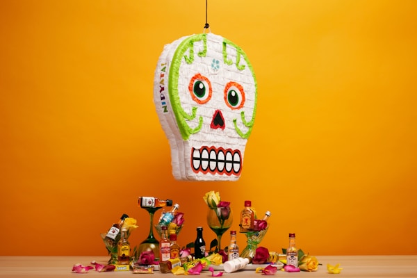 A colorful skull-shaped pi&ntilde;ata hangs against an orange backdrop. Below it is a wooden surface with various small liquor bottles, roses, and candy scattered around. The pi&ntilde;ata has bright green, orange, and red details, with a prominent toothy grin. The scene evokes a festive and lively atmosphere.