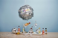 A large metallic piñata shaped like a disco ball hangs against a light blue background, with colorful letters spelling 'NIPYATA!' attached. Below, various miniature liquor bottles are arranged on a wooden surface, some in martini glasses. Smaller disco balls and confetti in star shapes add to the festive display.
