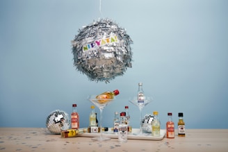 A large metallic piñata shaped like a disco ball hangs against a light blue background, with colorful letters spelling 'NIPYATA!' attached. Below, various miniature liquor bottles are arranged on a wooden surface, some in martini glasses. Smaller disco balls and confetti in star shapes add to the festive display.