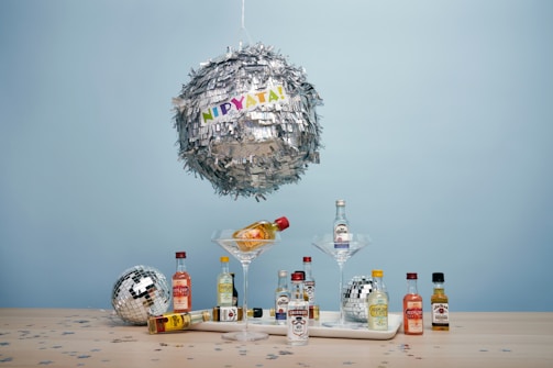A large metallic piñata shaped like a disco ball hangs against a light blue background, with colorful letters spelling 'NIPYATA!' attached. Below, various miniature liquor bottles are arranged on a wooden surface, some in martini glasses. Smaller disco balls and confetti in star shapes add to the festive display.