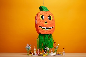 A colorful pumpkin-shaped piñata with a happy face hangs against a bright orange background. Below the piñata, various bottles of alcohol and snacks are arranged on a table. The surrounding contains green decorative elements, adding a festive touch.