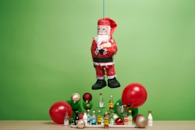 A Santa Claus piñata is suspended in front of a green backdrop. Below it, there is a wooden surface with an assortment of small liquor bottles and decorative items, including red and gold Christmas ornaments and balloons.