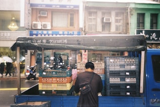 A man stands at the back of a blue truck loaded with stacks of plastic crates. The truck is parked on a street in front of a row of multi-story buildings with various signs in a foreign script. There are people walking on the sidewalk, some holding umbrellas, and a few scooters parked along the street.