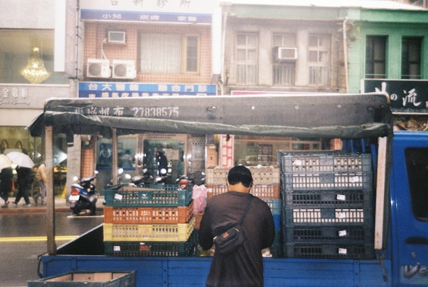 A man stands at the back of a blue truck loaded with stacks of plastic crates. The truck is parked on a street in front of a row of multi-story buildings with various signs in a foreign script. There are people walking on the sidewalk, some holding umbrellas, and a few scooters parked along the street.