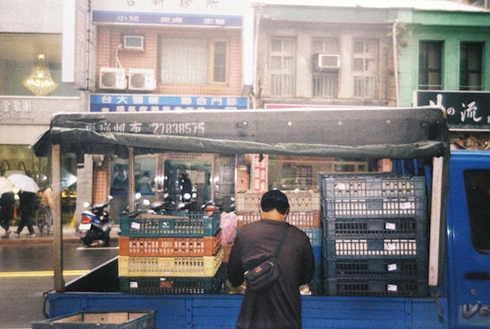 A man stands at the back of a blue truck loaded with stacks of plastic crates. The truck is parked on a street in front of a row of multi-story buildings with various signs in a foreign script. There are people walking on the sidewalk, some holding umbrellas, and a few scooters parked along the street.