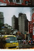 A bustling city street with a prominent yellow bus in the foreground, surrounded by tall buildings. People with umbrellas are walking on the sidewalks, and several scooters are on the road. There are visible storefronts including a KFC and various signs in different languages. The buildings display a mix of modern and traditional architecture.