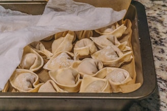 Several uncooked dumplings arranged in a baking tray, partially covered by a sheet of parchment paper. The dumplings appear to be made with a light, thin dough, and they are dusted lightly with flour. The tray is placed on a speckled countertop.