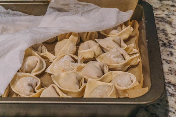 Several uncooked dumplings arranged in a baking tray, partially covered by a sheet of parchment paper. The dumplings appear to be made with a light, thin dough, and they are dusted lightly with flour. The tray is placed on a speckled countertop.