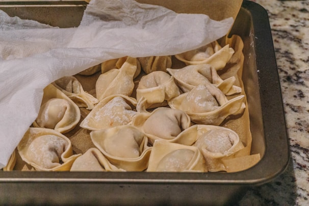 Several uncooked dumplings arranged in a baking tray, partially covered by a sheet of parchment paper. The dumplings appear to be made with a light, thin dough, and they are dusted lightly with flour. The tray is placed on a speckled countertop.