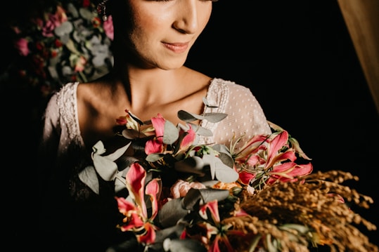 A woman is softly lit in warm tones, holding a bouquet with vibrant pink flowers and eucalyptus leaves. She is wearing a textured white garment with delicate embroidery. The overall composition is artful and intimate, capturing a tender moment.