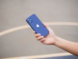 A hand is holding a blue smartphone with a prominent Apple logo on its back. The background is blurred, featuring a smooth, neutral-colored surface, possibly a concrete floor or playground.