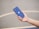 A hand is holding a blue smartphone with a prominent Apple logo on its back. The background is blurred, featuring a smooth, neutral-colored surface, possibly a concrete floor or playground.