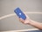 A hand is holding a blue smartphone with a prominent Apple logo on its back. The background is blurred, featuring a smooth, neutral-colored surface, possibly a concrete floor or playground.