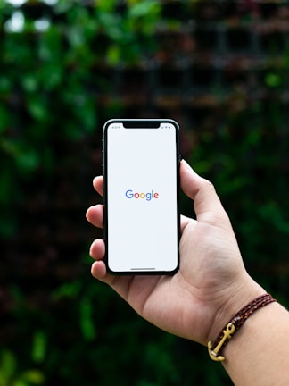 A hand holding a smartphone displaying the Google logo on the screen, set against a blurred natural background with greenery. The hand wears a brown and red braided bracelet adorned with a small metallic anchor charm.