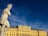 A classical stone statue of a warrior, possibly Greek or Roman, stands prominently with a contemplative posture. In the background, a grand yellow baroque-style palace is illuminated by the sunlight under a deep blue sky.