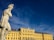 A classical stone statue of a warrior, possibly Greek or Roman, stands prominently with a contemplative posture. In the background, a grand yellow baroque-style palace is illuminated by the sunlight under a deep blue sky.