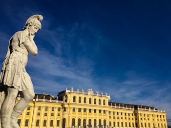 A classical stone statue of a warrior, possibly Greek or Roman, stands prominently with a contemplative posture. In the background, a grand yellow baroque-style palace is illuminated by the sunlight under a deep blue sky.