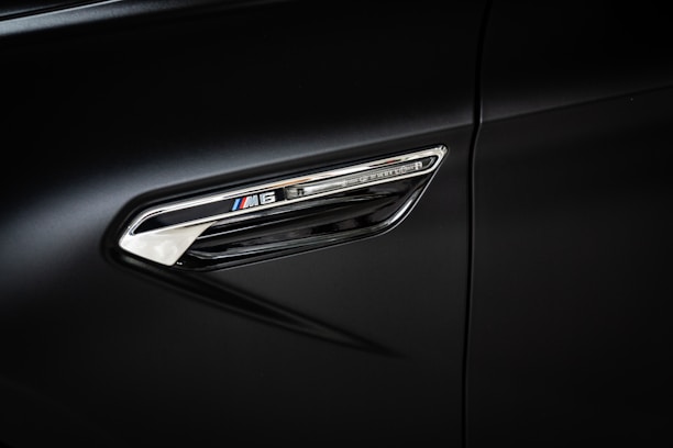 A close-up view of a car's side panel featuring a chrome detail with the logo and model designation 'M6'. The background is a sleek, dark surface that highlights the polished metal trim and its reflective quality.
