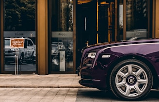A luxurious purple car is parked in front of a modern building with reflective glass doors. The building has the numbers '1699' displayed above the doors. A sign with Asian characters is visible near the entrance. The scene conveys a sense of opulence and urban sophistication.