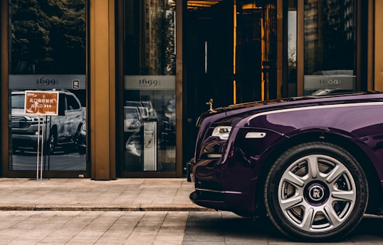 A luxurious purple car is parked in front of a modern building with reflective glass doors. The building has the numbers '1699' displayed above the doors. A sign with Asian characters is visible near the entrance. The scene conveys a sense of opulence and urban sophistication.