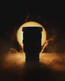 A silhouetted camera lens is placed against a bright circular light source, creating a dramatic backlight effect. Wisps of smoke or mist surround the lens, adding an enigmatic atmosphere to the scene.