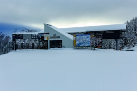A snow-covered ski lift station is situated in a mountainous region, with snow blanketing the ground. The building has a sloped roof and displays signage, including large maps and instructions. The surrounding trees and mountains create a wintery backdrop with dense clouds in the sky, suggesting overcast weather.