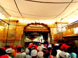 A large crowd of people wearing vibrant turbans is gathered in front of an ornate, golden entrance, likely part of a religious or cultural site. Decorative carvings and script adorn the entrance, and the area is shaded by a large canopy.