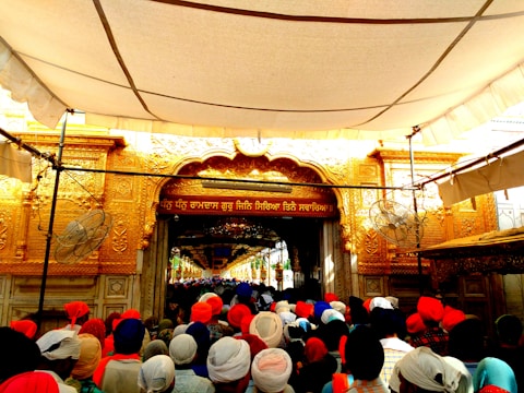 A large crowd of people wearing vibrant turbans is gathered in front of an ornate, golden entrance, likely part of a religious or cultural site. Decorative carvings and script adorn the entrance, and the area is shaded by a large canopy.