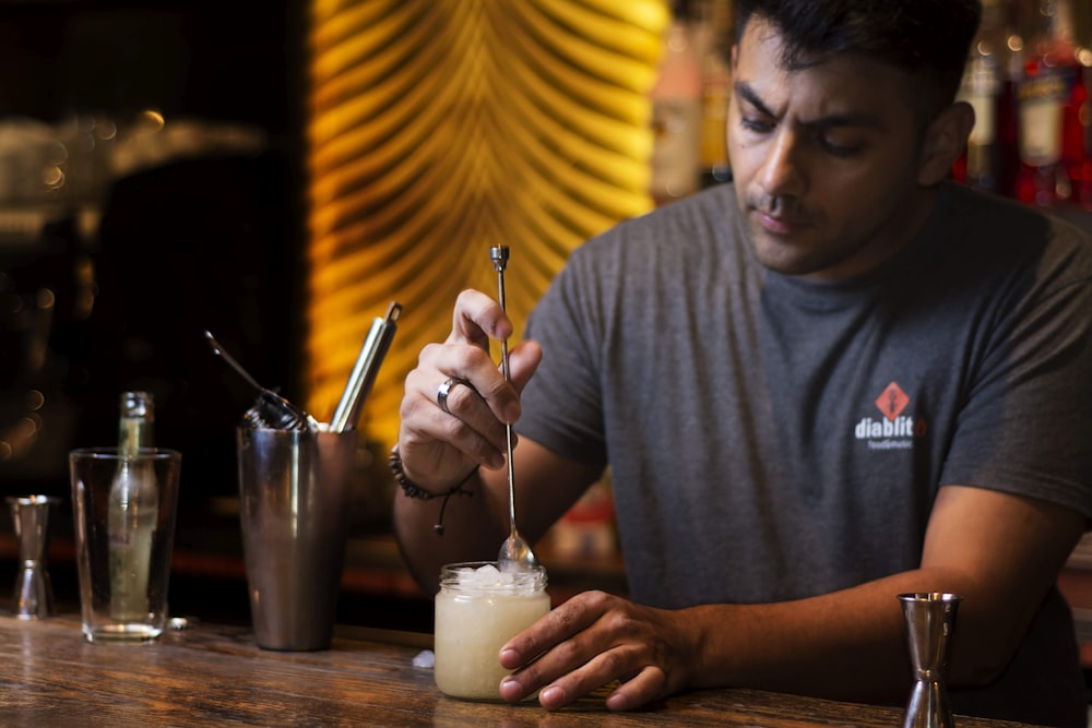 homem na camisa cinza do pescoço da tripulação segurando caneta prateada e líquido branco no copo de beber transparente