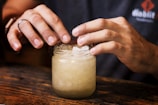 Hands are carefully placing crushed ice into a glass jar. The jar is placed on a wooden surface, and the person is wearing a dark shirt bearing a visible logo on the sleeve. The lighting emphasizes the textures on the hands and the ice.