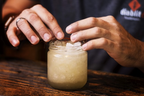 Hands are carefully placing crushed ice into a glass jar. The jar is placed on a wooden surface, and the person is wearing a dark shirt bearing a visible logo on the sleeve. The lighting emphasizes the textures on the hands and the ice.