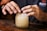 Hands are carefully placing crushed ice into a glass jar. The jar is placed on a wooden surface, and the person is wearing a dark shirt bearing a visible logo on the sleeve. The lighting emphasizes the textures on the hands and the ice.
