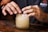 Hands are carefully placing crushed ice into a glass jar. The jar is placed on a wooden surface, and the person is wearing a dark shirt bearing a visible logo on the sleeve. The lighting emphasizes the textures on the hands and the ice.
