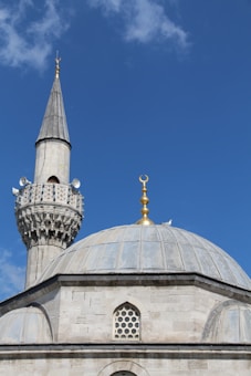 A mosque featuring a prominent dome and a tall minaret with intricate architectural details stands against a clear blue sky. The dome is topped with a golden crescent finial, and the minaret is adorned with decorative motifs and loudspeakers.