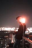 A person in a hooded jacket stands on a rooftop at night, holding a camera. They are leaning against a railing, with a red light illuminating their face. The city skyline is visible in the background, with numerous lights softly glowing.