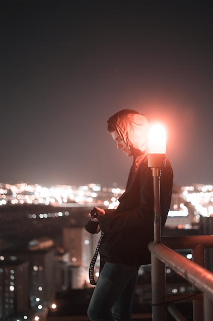 A person in a hooded jacket stands on a rooftop at night, holding a camera. They are leaning against a railing, with a red light illuminating their face. The city skyline is visible in the background, with numerous lights softly glowing.