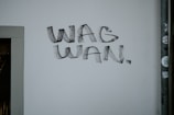 A wall with the phrase 'WAG WAN' written in bold, black letters. The wall is mostly plain and there is a vertical pipe with some small stickers on it to the right.