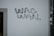A wall with the phrase 'WAG WAN' written in bold, black letters. The wall is mostly plain and there is a vertical pipe with some small stickers on it to the right.