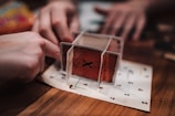 A close-up view of two hands interacting with a structure made of cards on a wooden table. The structure appears somewhat unstable and creative, using cards with numbers and letters printed on them, possibly for a game or puzzle.