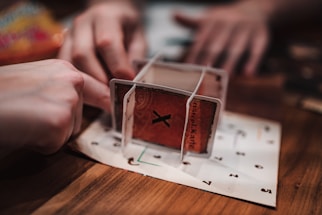 A close-up view of two hands interacting with a structure made of cards on a wooden table. The structure appears somewhat unstable and creative, using cards with numbers and letters printed on them, possibly for a game or puzzle.