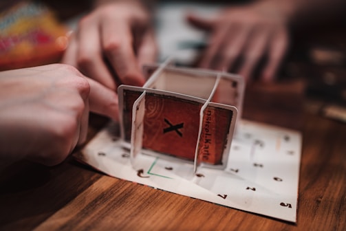 A close-up view of two hands interacting with a structure made of cards on a wooden table. The structure appears somewhat unstable and creative, using cards with numbers and letters printed on them, possibly for a game or puzzle.