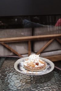 A dessert placed on a reflective glass table features a round pastry topped with a scoop of ice cream, caramelized nuts, and a sprinkling of powdered sugar. The background includes a cushioned bench with a wooden frame, creating a cozy café setting.