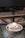 A dessert placed on a reflective glass table features a round pastry topped with a scoop of ice cream, caramelized nuts, and a sprinkling of powdered sugar. The background includes a cushioned bench with a wooden frame, creating a cozy café setting.