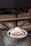 A dessert placed on a reflective glass table features a round pastry topped with a scoop of ice cream, caramelized nuts, and a sprinkling of powdered sugar. The background includes a cushioned bench with a wooden frame, creating a cozy café setting.