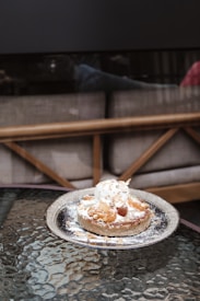 A dessert placed on a reflective glass table features a round pastry topped with a scoop of ice cream, caramelized nuts, and a sprinkling of powdered sugar. The background includes a cushioned bench with a wooden frame, creating a cozy caf&eacute; setting.