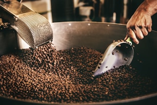 A person is using a metal scoop to handle roasted coffee beans that are being poured into a large container. The atmosphere is warm and industrial, suggesting a coffee roasting process.