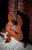 A classical acoustic guitar is leaning against a brick wall. Below it, there are music sheets scattered around, adorned with autumn leaves. The setting conveys a rustic and artistic ambiance.