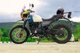 A Royal Enfield Himalayan motorcycle is parked on a paved area with a scenic backdrop of hills and greenery. The motorcycle is equipped with a helmet placed on the rear. It features a tank adorned with a colorful garland. The sky is overcast, suggesting a cloudy day.