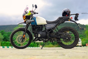 A Royal Enfield Himalayan motorcycle is parked on a paved area with a scenic backdrop of hills and greenery. The motorcycle is equipped with a helmet placed on the rear. It features a tank adorned with a colorful garland. The sky is overcast, suggesting a cloudy day.